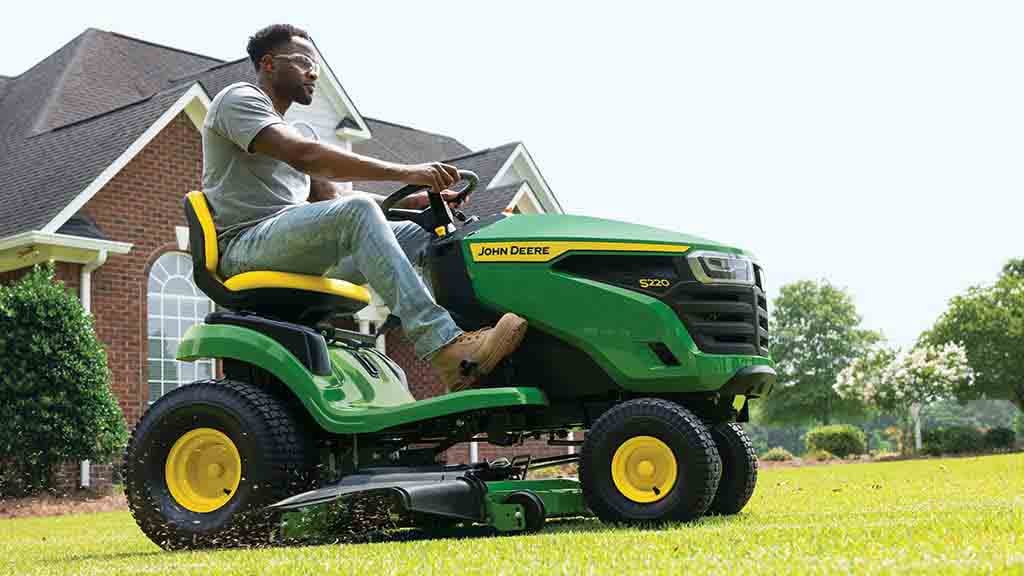 Gros plan d’une personne conduisant une tondeuse autoportée verte avec des coupures d’herbe volant hors du plateau.
