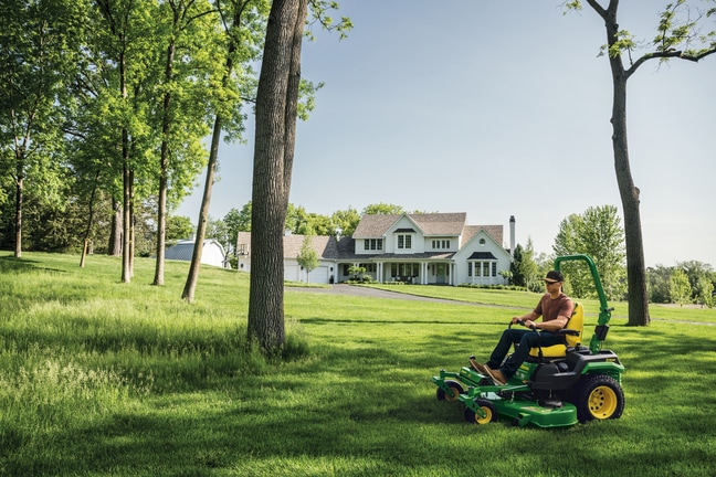 A green zero-turn mower is heading towards some tall grass growing between spread-out trees.