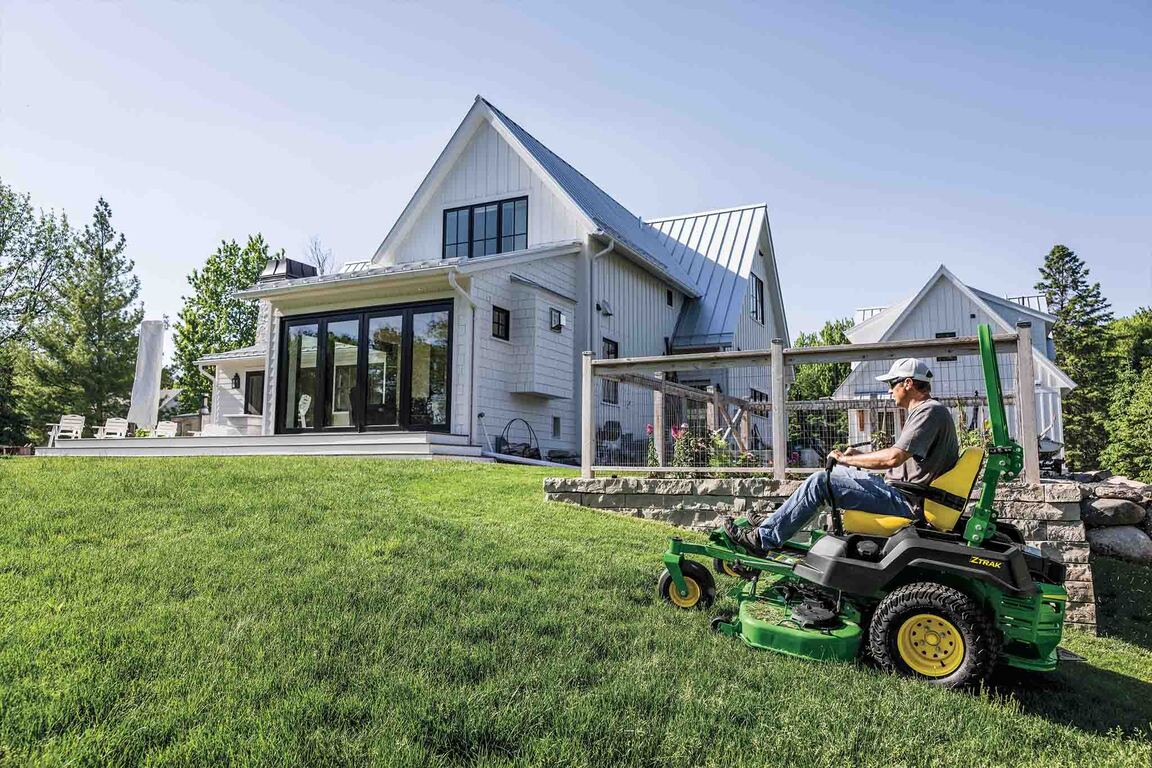 A green zero-turn mower is mowing up a gentle slope near a fenced-off garden.