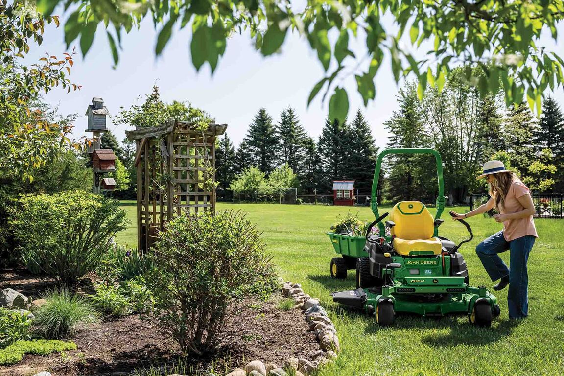 A person steps onto a zero-turn mower with a poly cart attachment hauling plants.