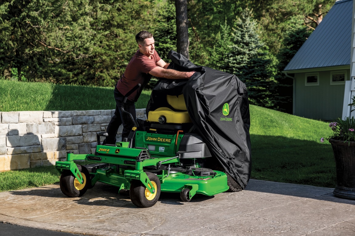 A cover with the Deere logo being removed from a green zero-turn mower in a driveway.