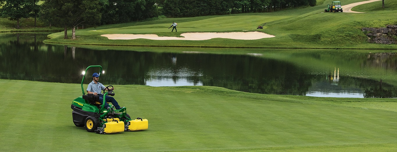 Person mowing golf course with pond, sand traps, and another person in the background.