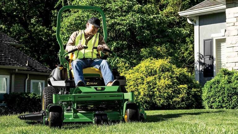 Grass being mowed in front of a residence by a landscaper on a green zero-turn mower.