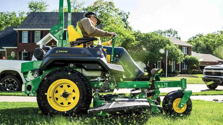 A landscaper operating a green zero-turn mower in a residential area with blades of cut grass coming towards the camera.
