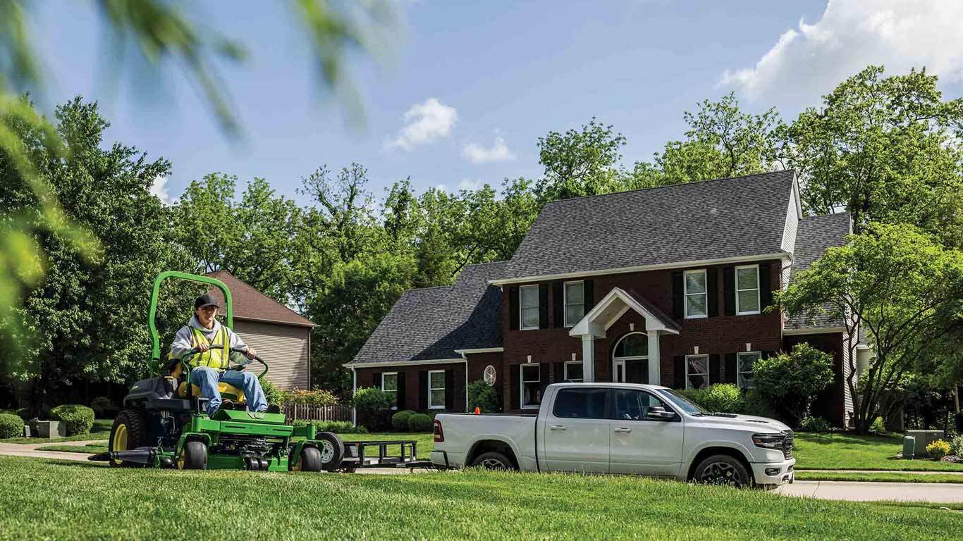 A landscaper driving a green zero-turn mower by a trailer in a residential area.