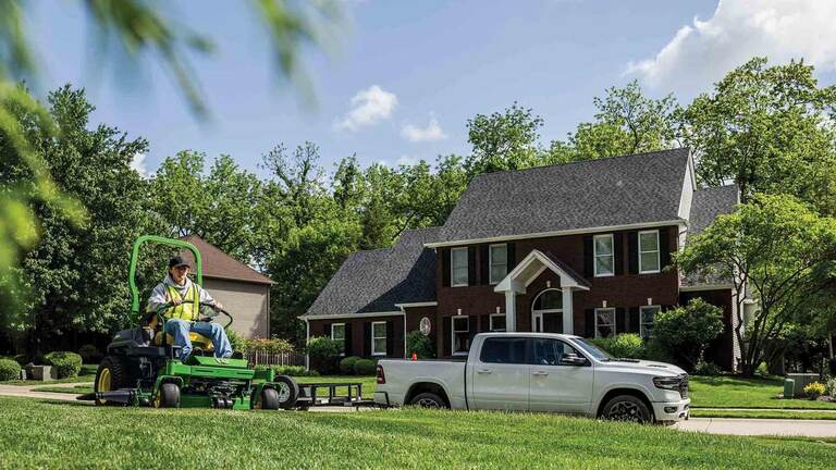 A landscaper driving a green zero-turn mower by a trailer in a residential area.