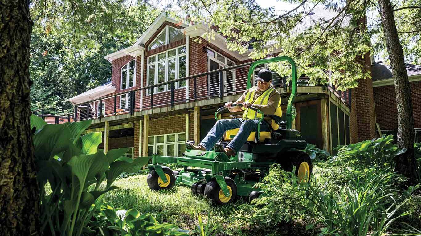 A landscaper mowing grass along the edge of a garden behind a residence with a green zero-turn mower.