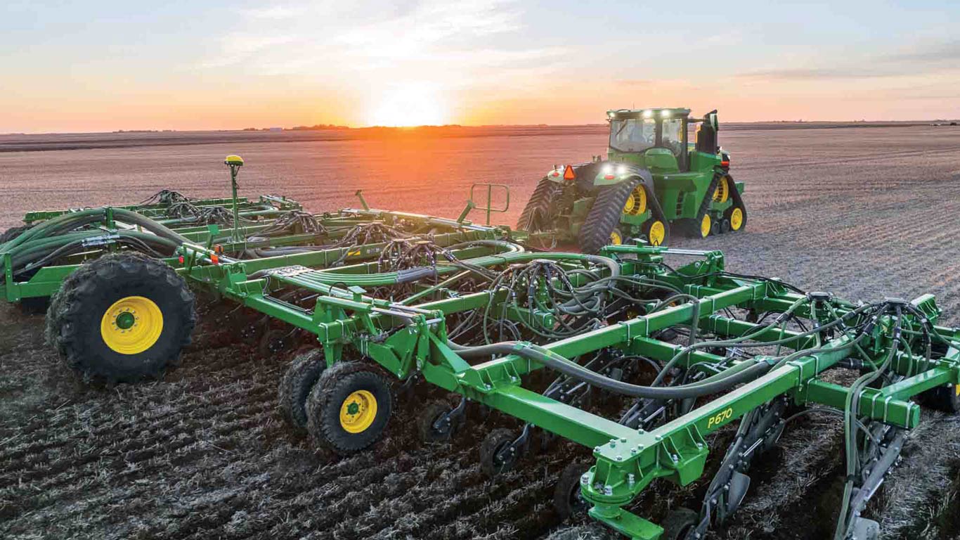 Vue latérale arrière d’un semoir à houes pneumatique de précision P670 tracté dans un champ par un tracteur John Deere chenillé de la série&nbsp;8, avec le lever du soleil en arrière-plan.