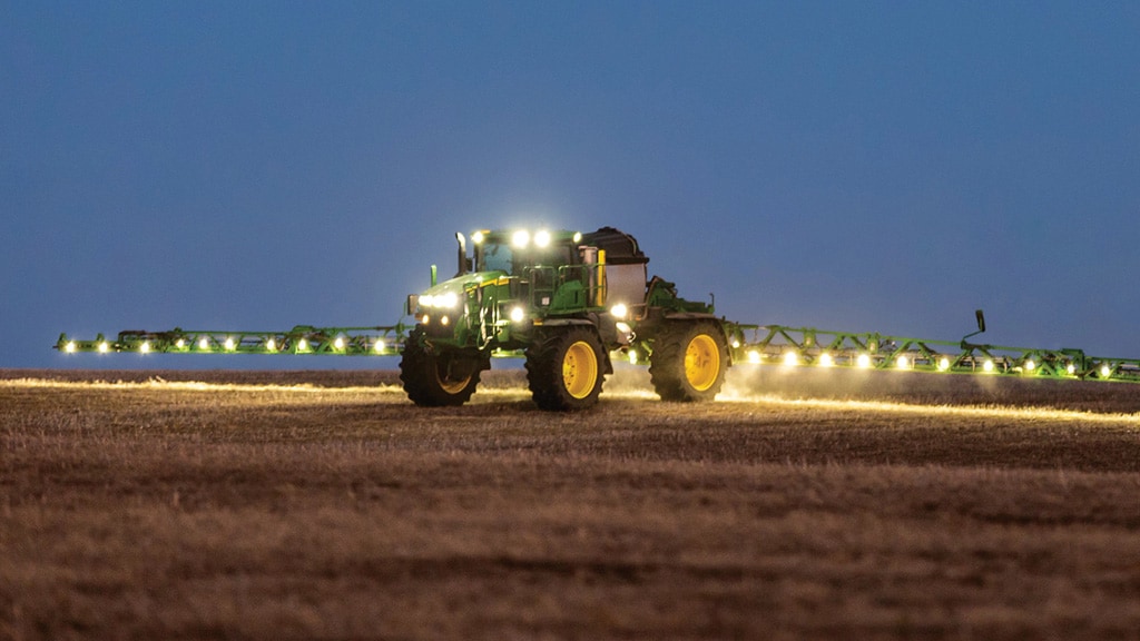 Photo showing a John Deere Sprayer making a dusk application pass with fully extended, lighted booms.