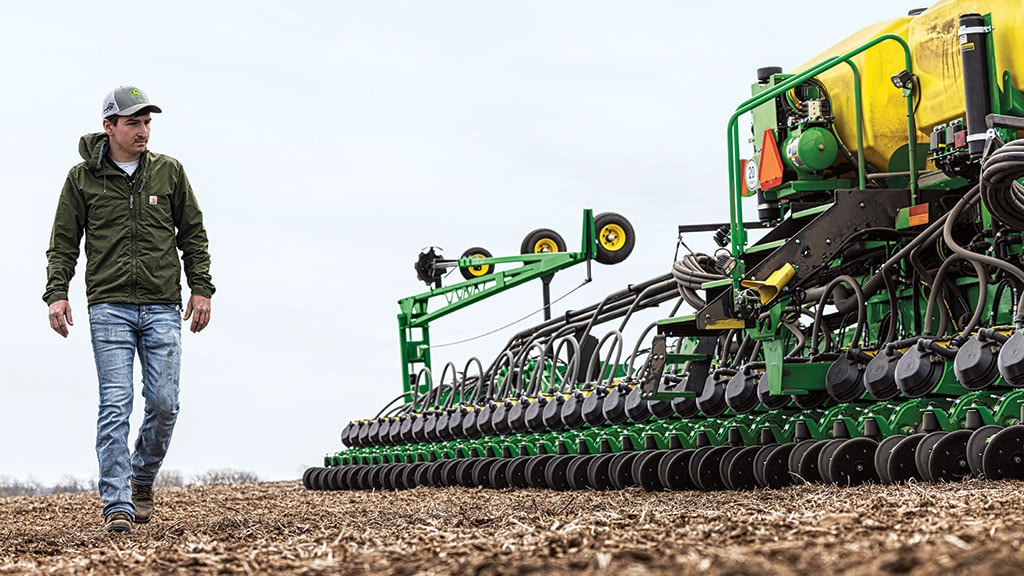 Une personne marche à côté de machines agricoles dans un champ, avec des rangées d’équipement d’ensemencement et un ciel nuageux en arrière-plan.