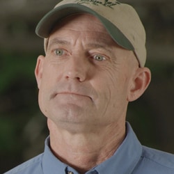 Un agriculteur portant une casquette et une chemise claire boutonnée, sur fond sombre.