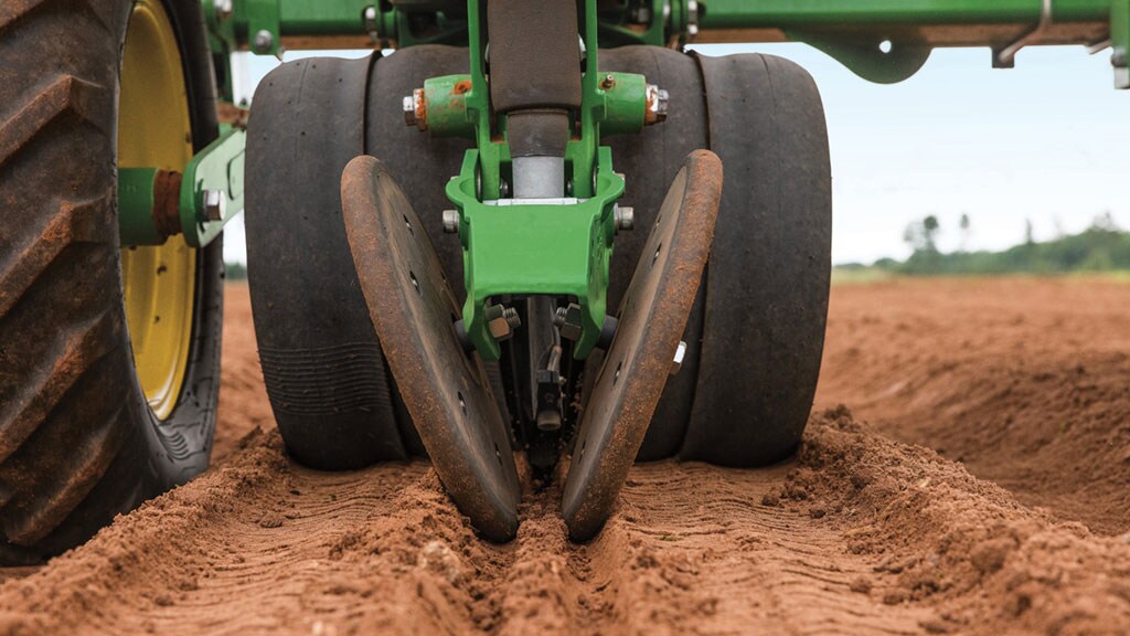 Ground-level close-up of John Deere planter double-disc openers cutting into red soil to create a seed furrow, with gauge wheels and tractor tires visible.