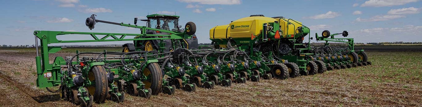 A green tractor pulling a wide central-fill planter with a yellow commodity tank across a field under a bright blue sky with scattered clouds.