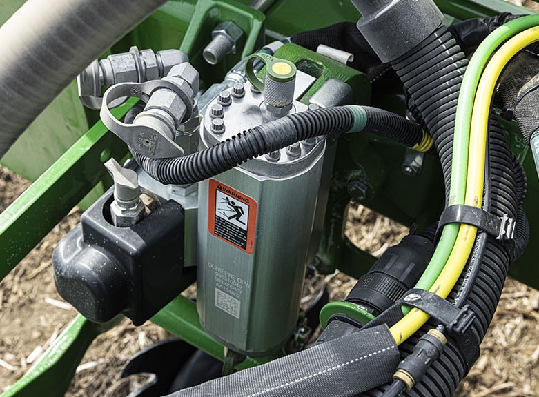 Vue en gros plan d’un vérin hydraulique sur un châssis de semoir de précision John Deere, entouré de flexibles et de câbles, avec un autocollant l’avertissement rouge apposé.