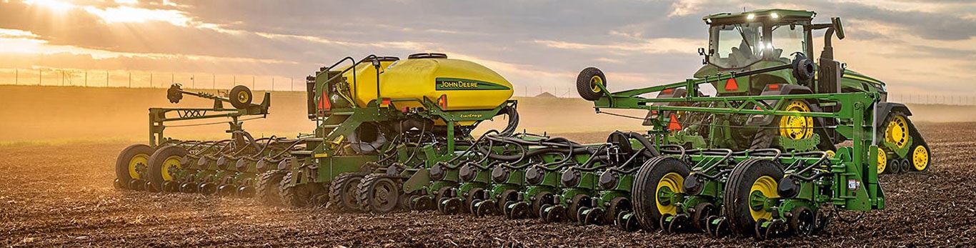 A John Deere tractor pulling an ExactEmerge planter across a field at sunset, with golden light and dust trailing in the background.