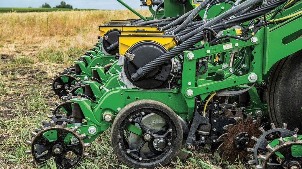 Gros plan des éléments à rangs du semoir de précision John Deere dans un champ, montrant les roues de couvre-sillons, les accessoires de nettoyage de rangs et les tubes à semences.