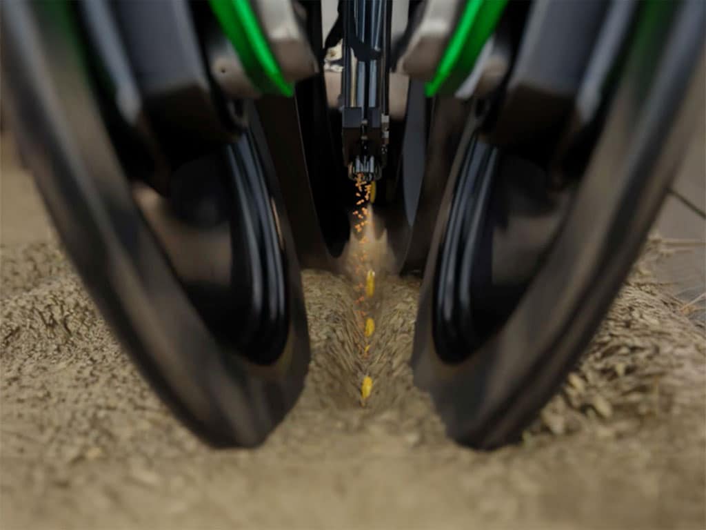 Gros plan sur un semoir déposant des graines dans le sol entre deux roues noires.