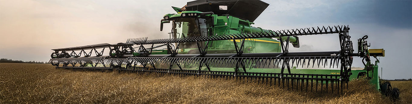 A green John Deere combine harvester with extended header cutting through a golden grain field under a clear sky