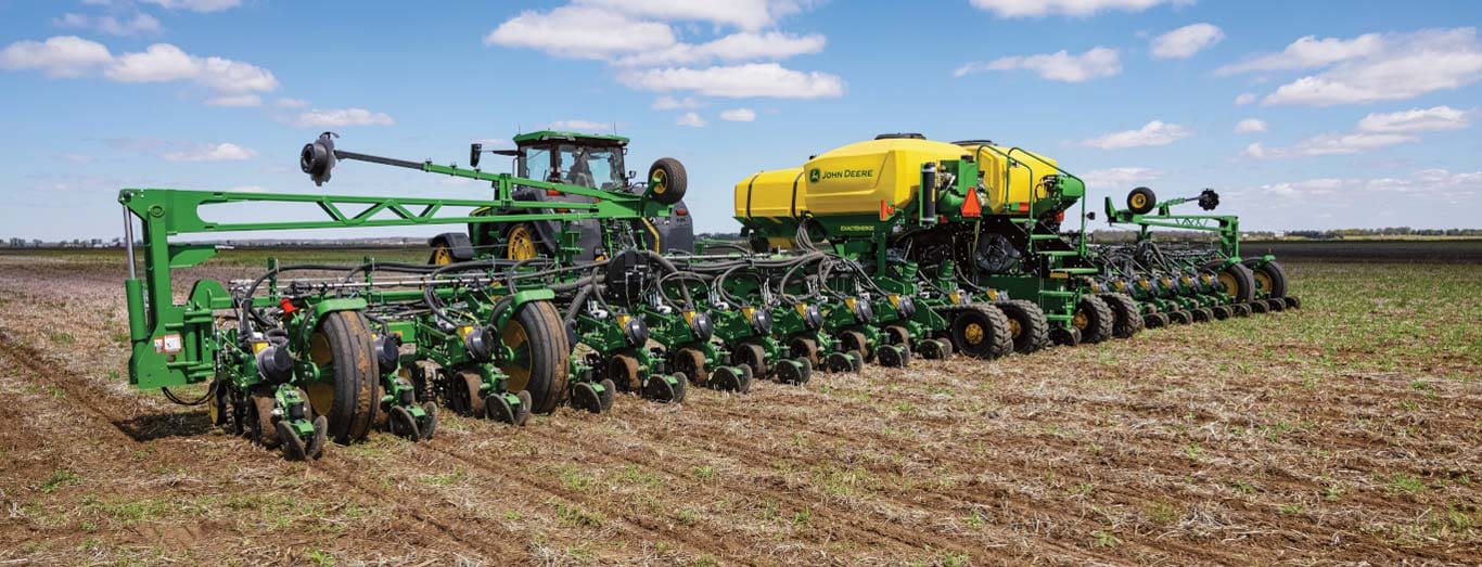 Un tracteur vert tirant un semoir de précision large à remplissage central avec un réservoir de produit jaune à travers un champ sous un ciel bleu vif parsemé de nuages.