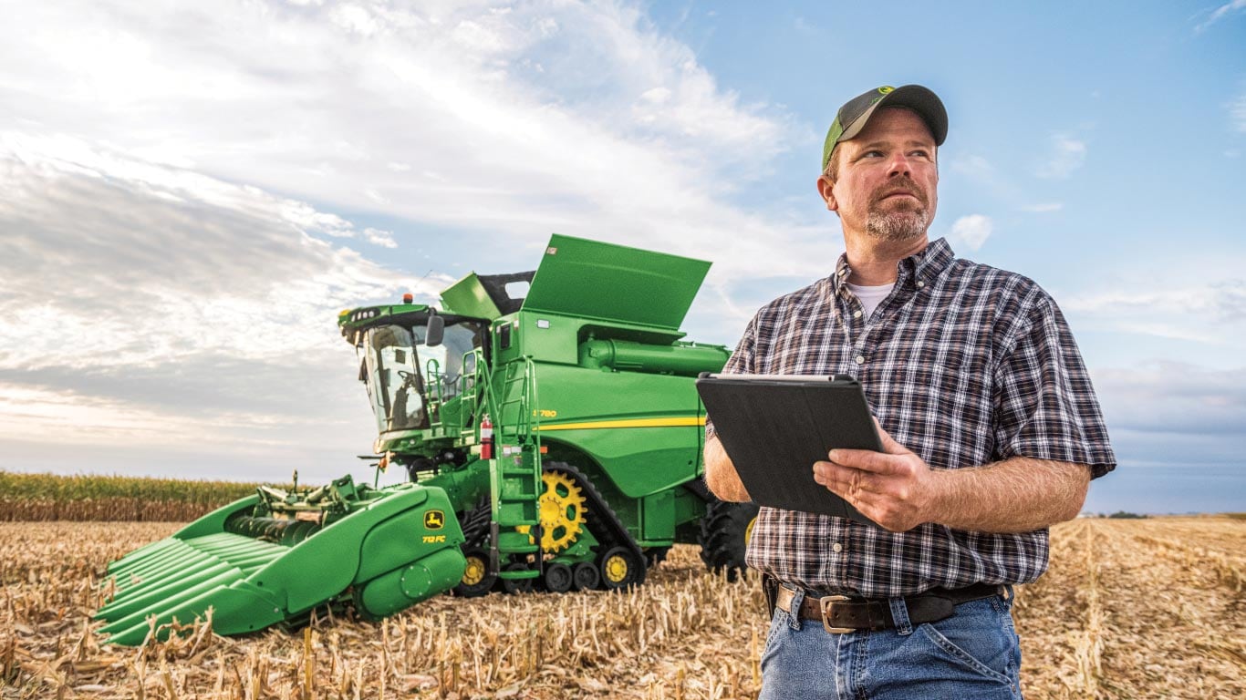 Un agriculteur vêtu d’une chemise à carreaux et d’une casquette, tablette à la main, devant une moissonneuse-batteuse verte dans un champ.