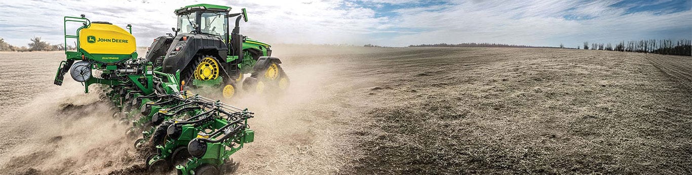 John Deere tractor pulling an ExactEmerge planter across a large open field, with dust trailing behind under a partly cloudy sky.