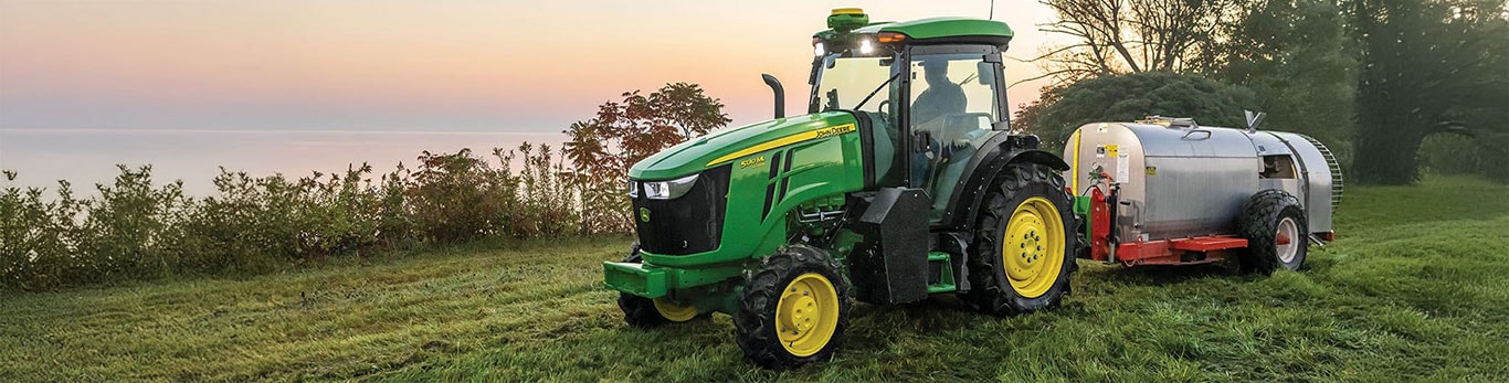 A John Deere 5100M tractor with a silver liquid sprayer tank parked on a grassy hillside at dusk, with trees and water visible in the background.