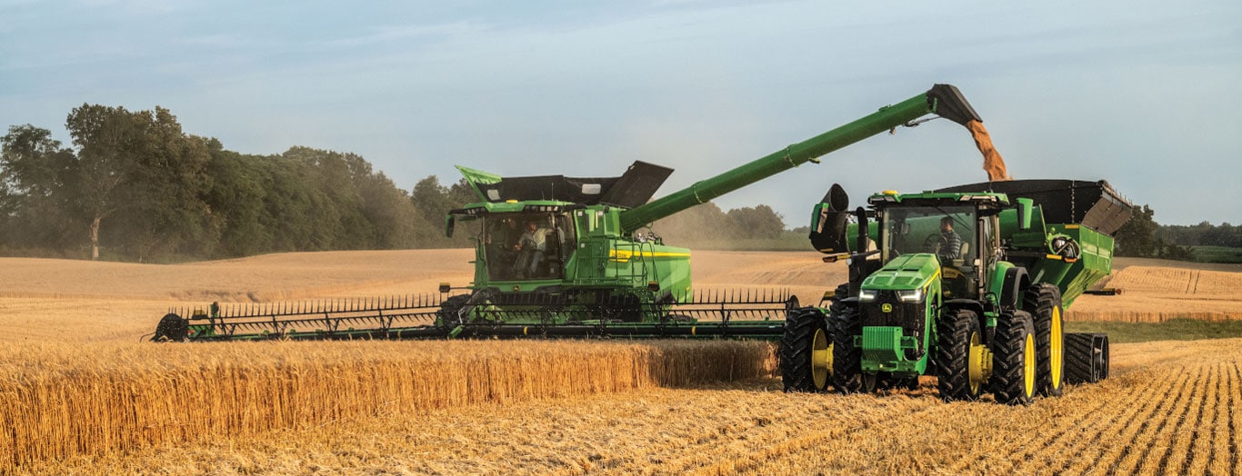 Une moissonneuse-batteuse verte récolte du blé et décharge le grain à la volée dans une remorque tirée par un tracteur, au milieu d’un champ doré.