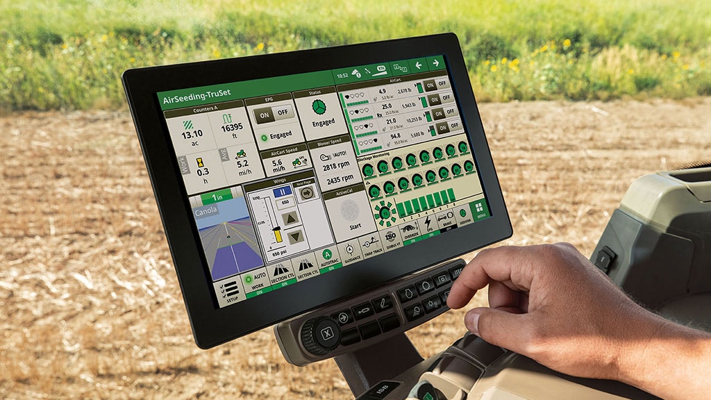 Farmer's hand holding a tablet displaying AirSeeding TruSet precision agriculture software with crop data, field maps, and equipment monitoring screens visible on the display, with a harvested grain field in the background