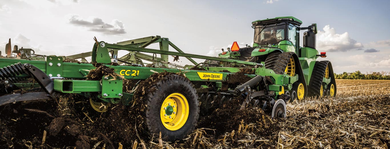 Un tracteur tirant un outil de labourage du sol à travers d’importants résidus de maïs selon un angle faible, retournant la terre sous une lumière dorée.