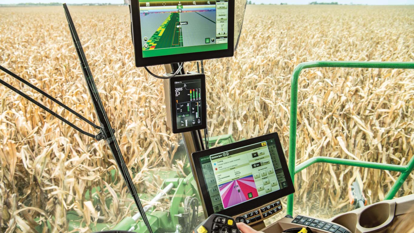 Vue dans la cabine des consoles John Deere installées dans une moissonneuse-batteuse, avec la main de l’utilisateur sur le levier de commande et du maïs à maturité visible à travers la vitre.