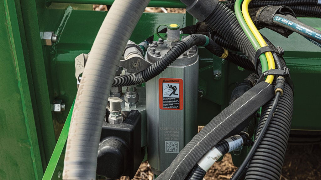 Close-up of a hydraulic cylinder on a John Deere planter frame, surrounded by hoses and cables, with a red warning label attached.