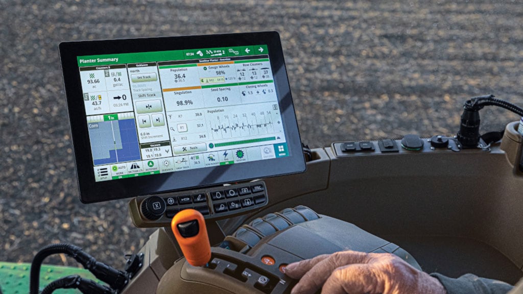 Image de la main de la personne sur un joystick dans une cabine de tracteur, avec une tablette affichant l’écran récapitulatif du semoir de précision.