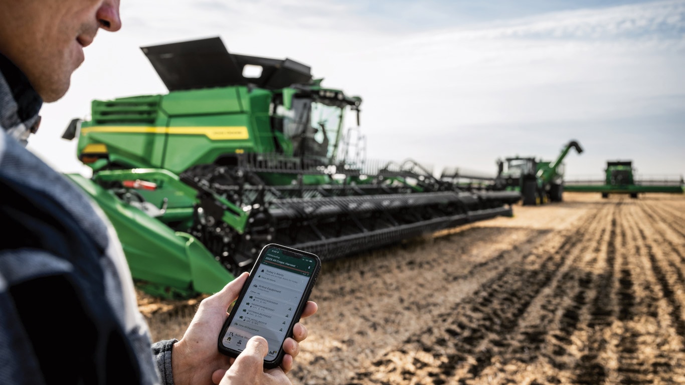 Une personne consultant l’application John Deere Equipment Mobile sur un téléphone intelligent tandis qu’une moissonneuse-batteuse récolte derrière elle.