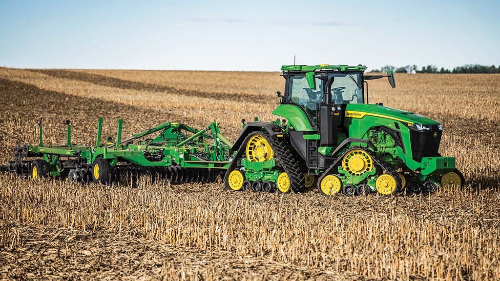 Tracteur vert tirant un équipement de labourage dans un champ de chaume récolté, sous un ciel bleu.