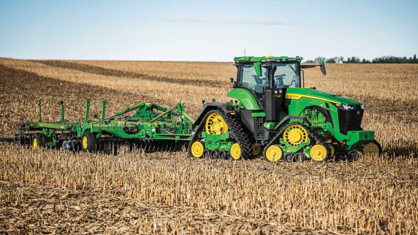 Tracteur vert John Deere tirant un équipement de labourage dans un champ de chaume récolté, sous un ciel bleu.