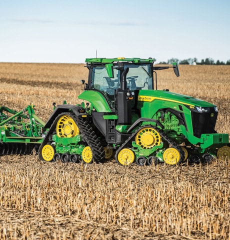 Tracteur vert tirant un équipement de labourage dans un champ de chaume récolté, sous un ciel bleu.