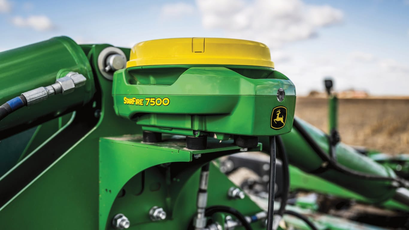 Un récepteur StarFire™ 7500 de John Deere, un module de guidage vert au capot jaune, monté sur un équipement devant un ciel nuageux.