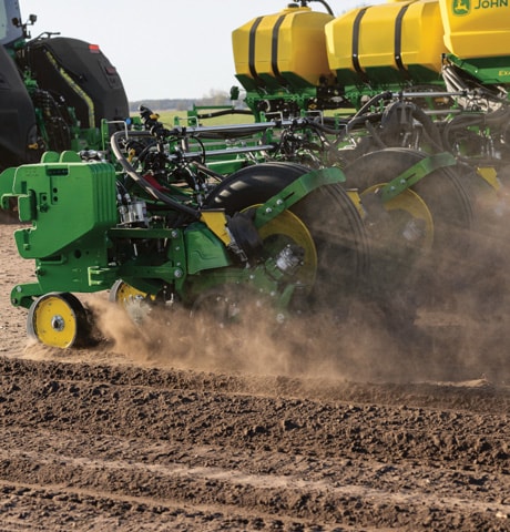 Tracteur vert tirant un semoir de précision à travers un sol labouré, soulevant la poussière.