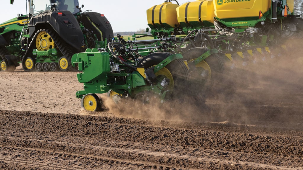 Tracteur vert tirant un semoir de précision à travers un sol labouré, soulevant la poussière.
