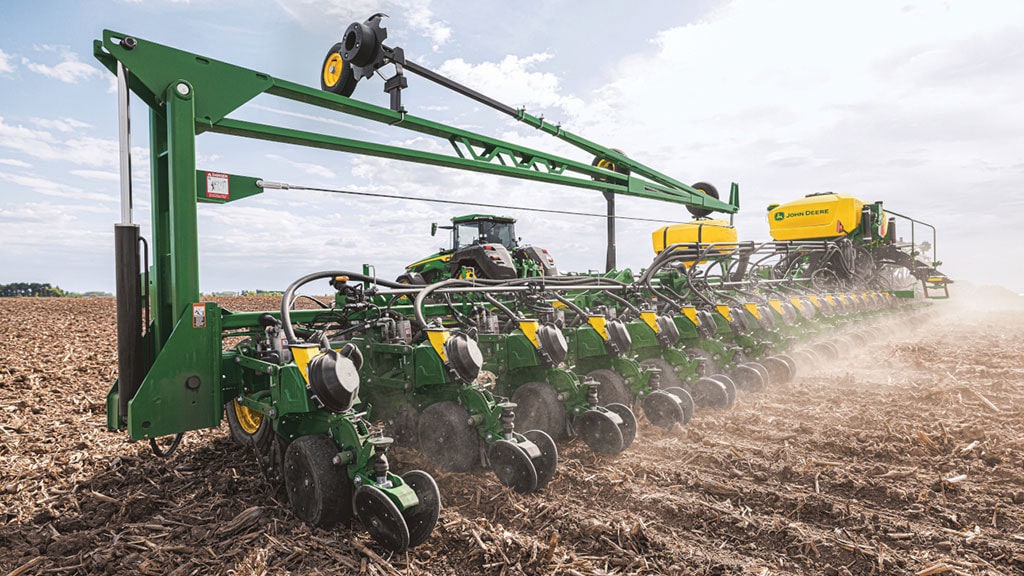 Semoir de précision à rangs multiples John Deere utilisé sur un champ de résidus de récolte, avec un tracteur visible en arrière-plan.