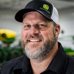 Un agriculteur, coiffé d’une casquette et vêtu d’une chemise foncée, sourit dans un atelier devant un semoir de précision John Deere vert.