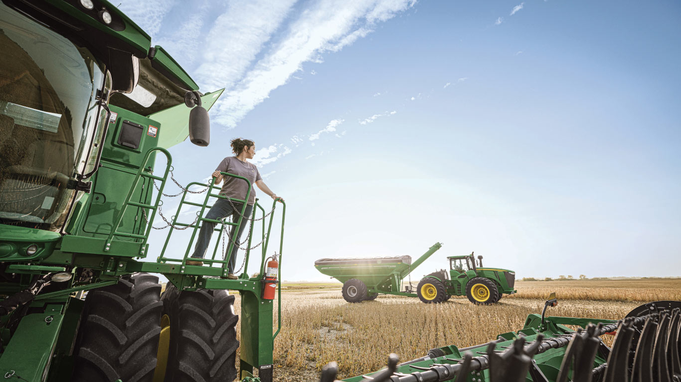 Sur la plate-forme arrière d’une moissonneuse-batteuse verte, une utilisatrice observe un chariot à grain et un tracteur dans un champ de blé, sous un ciel bleu.