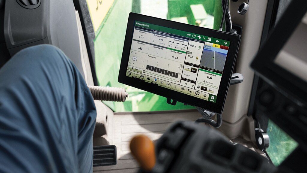 Tractor cab display showing a John Deere SeedStar planting interface with population, singulation, seed spacing, and row cleaner data, with a farmer visible in the seat.