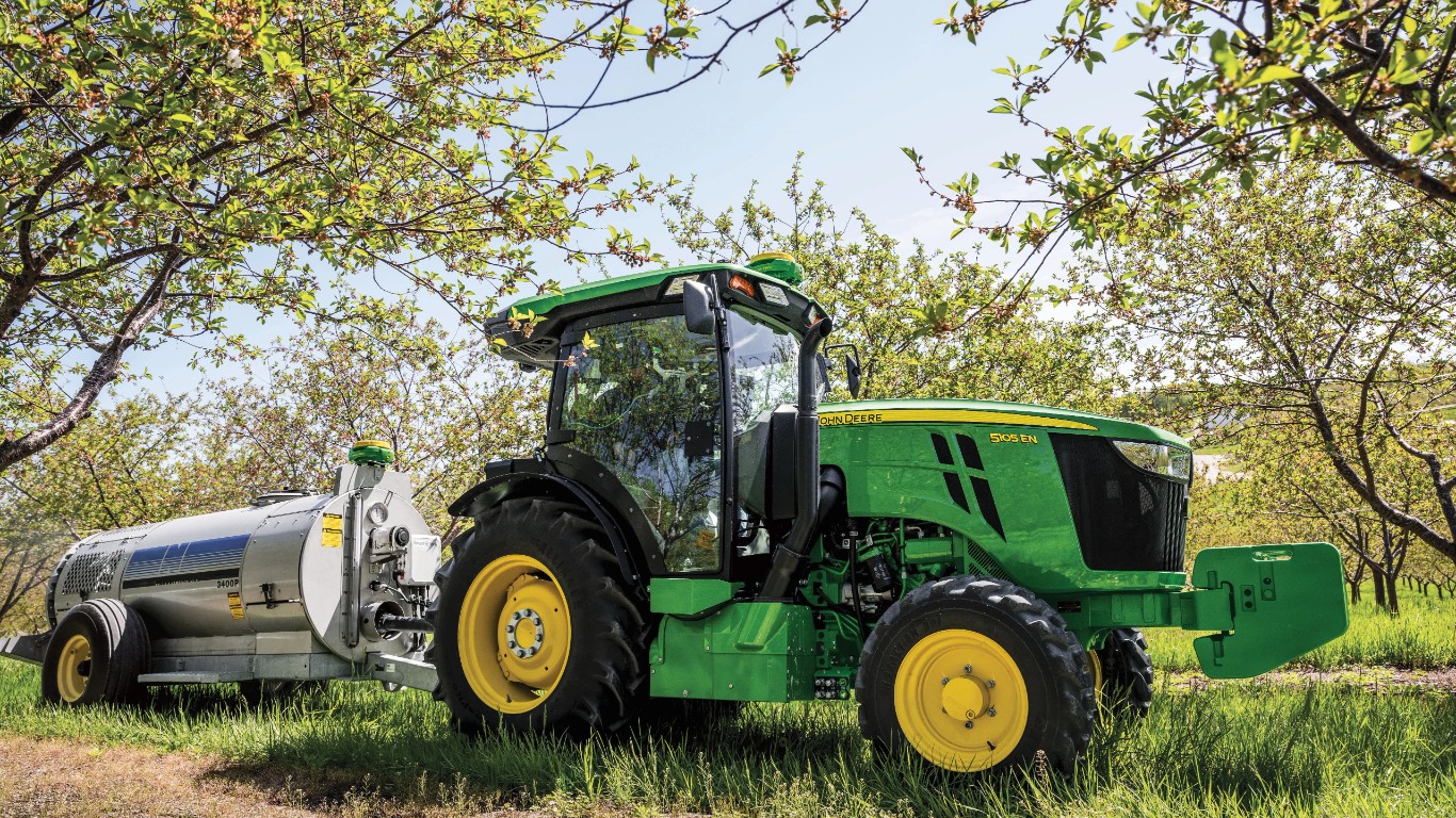 A John Deere 5105EN tractor with a silver liquid applicator tank parked in an orchard under leafy trees.