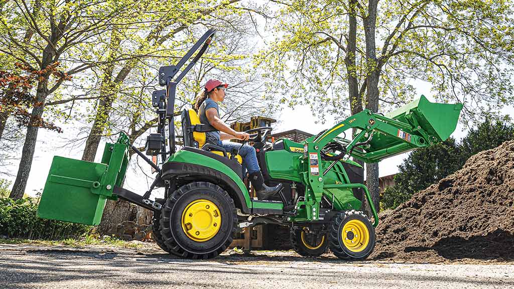 A green compact tractor with a front loader and rear ballast box scooping up soil.