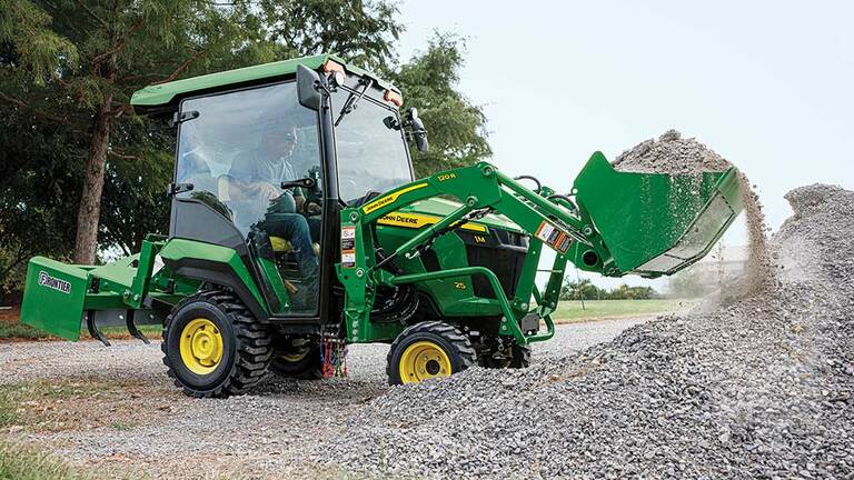 A green compact tractor with a cab, land plane, and loader scooping up gravel.
