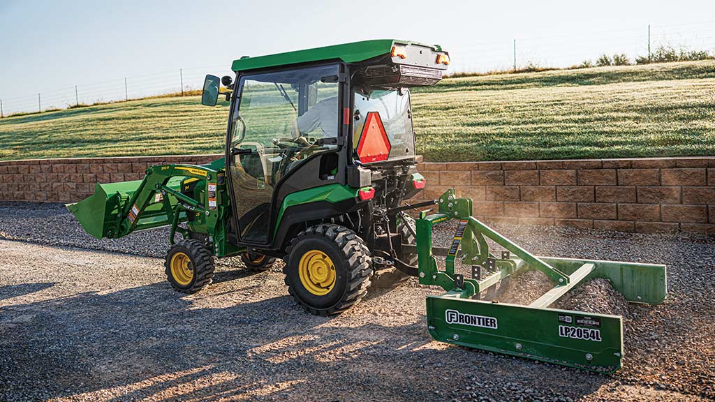A cabbed green compact tractor with a front loader and a rear land plane leveling gravel next to a retaining wall.