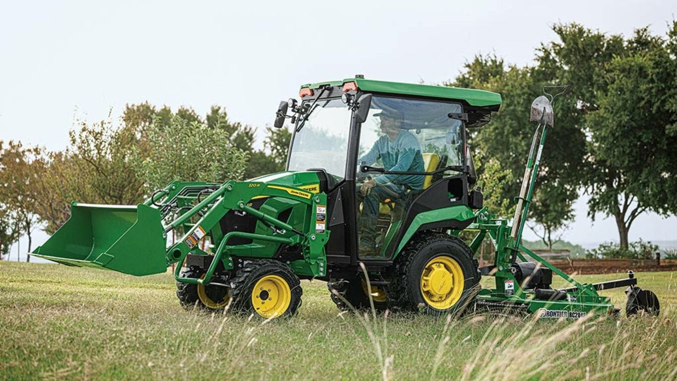A cabbed green compact tractor with a loader pulls a rotary cutter over some grass.