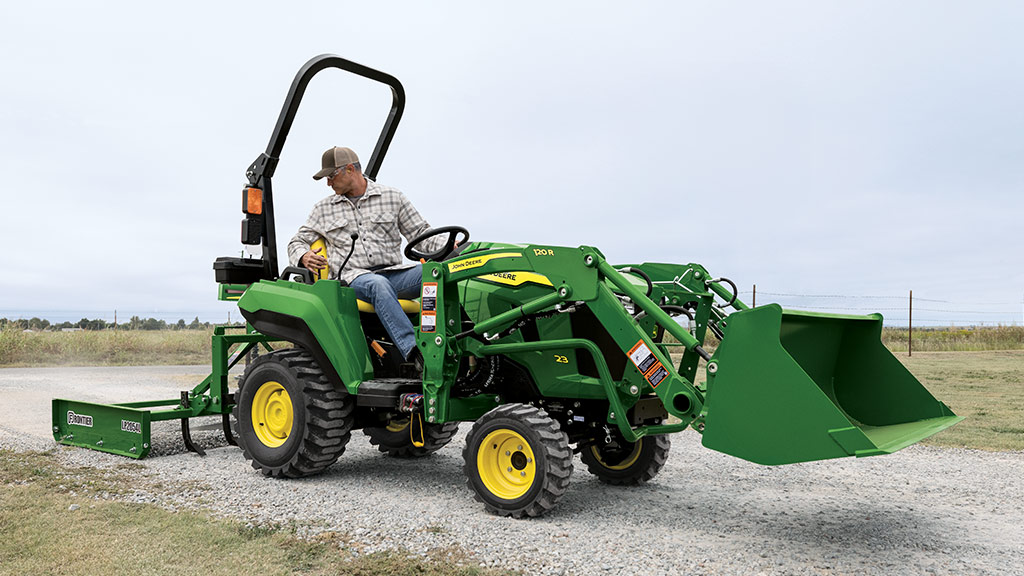 A person driving a green compact tractor equipped with a loader and land plane spreading gravel on a gravel road.