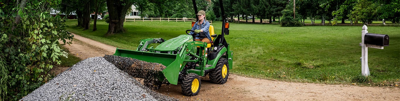 A person in a green compact tractor on a dirt path scooping up gravel with an attached front loader.